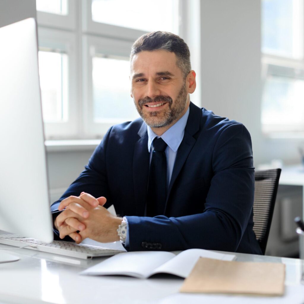 Portrait of happy middle aged businessman sitting in front of computer at workplace in office, smiling to camera. Entrepreneur man sitting at desk. Business and entrepreneurship